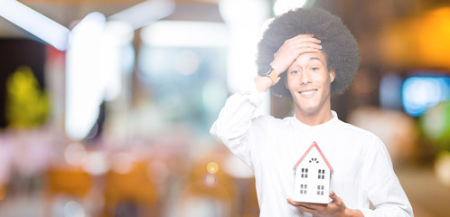 Young african american man with afro hair holding house stressed with hand on head, shocked with...