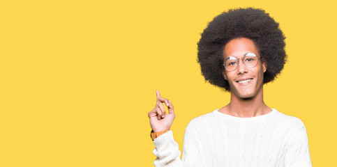 Young african american man with afro hair wearing glasses with a big smile on face, pointing with hand and finger to the side looking at the camera.