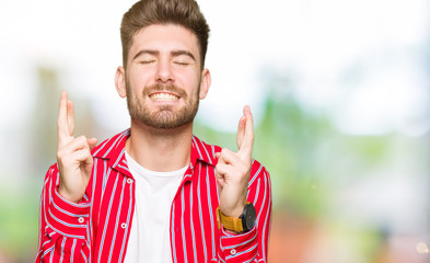 Young handsome man wearing red shirt smiling crossing fingers with hope and eyes closed. Luck and superstitious concept.