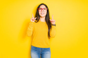 Beautiful brunette woman wearing red glasses over yellow isolated background amazed and surprised looking up and pointing with fingers and raised arms.
