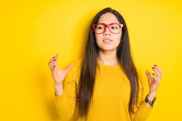 Beautiful brunette woman wearing red glasses over yellow isolated background crazy and mad shouting and yelling with aggressive expression and arms raised. Frustration concept.