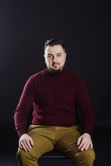 Fototapeta na wymiar a man with a beard in casual clothes poses on an isolated dark won, in a burgundy sweater in the studio