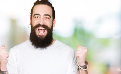 Young hipster man with long hair and beard wearing casual white t-shirt celebrating surprised and amazed for success with arms raised and open eyes. Winner concept.