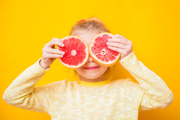 Smiling child girl holding two halfs of yellow grapefruit citrus fruit in hands, covering her eyes, on yellow. Healthy diet nutrition. Happiness fun concept