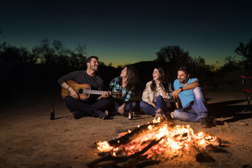 Group of friends drinking and singing next to bonfire while camping at night