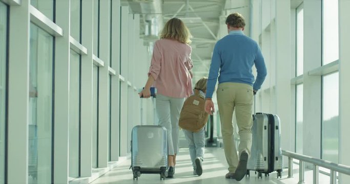 Rear of the Caucasian small boy walking to the arrival gates in the airport corridor and his parents following with suitcases on the wheels.