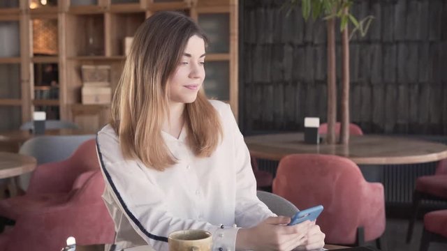 A pretty girl enjoys herself in restaurant during lunch hour. She uses her phones, turns her favourite music and keeps dancing while sitting at the table. The lady is happy. The background is blurred