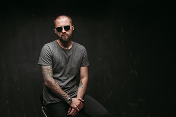 Portrait of a handsome balded man with long well trimmed beard wearing sunglasses and grey shirt looking at camera.