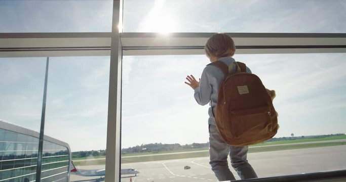 Back view on the small cute Caucasian boy with a backpack standing at the big glass window in the airport and looking outside at the pitch. Rear.
