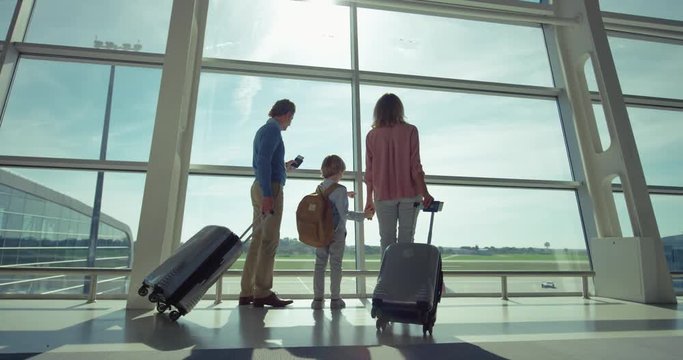 Rear of the caucasian parents with suitcases on wheels and little son with a backpack standing at the big glass window in the airport and looking outside. Back view.