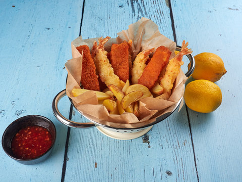 Fried fish fingers, tempura prawns and potato wedges in a colander, with lemons and sriracha sauce, on blue wooden background