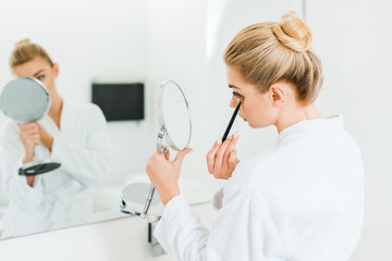Obraz premium selective focus of woman in white bathrobe applying eyeshadow with cosmetic brush