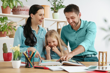 Fototapeta premium Father, mother and their daughter are smiling while spending time together. A day with family. Young happy couple with child are studying with the book. Education, studying and knowledge sharing