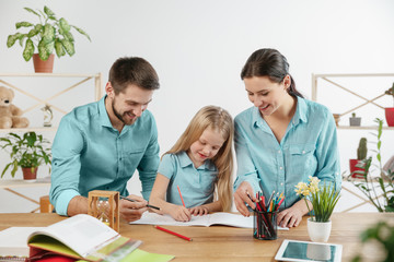Father, mother and their daughter are smiling while spending time together. A day with family. Young happy couple with child are learning to draw. Education, studying and knowledge sharing concept.