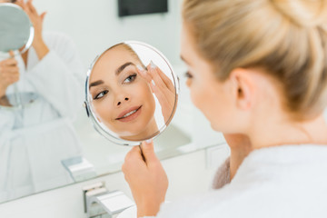 selective focus of attractive woman touching face and looking at mirror in bathroom