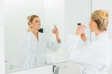 selective focus of beautiful woman in white bathrobe brushing teeth and taking selfie in bathroom