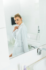 beautiful and smiling woman in white bathrobe looking at mirror in bathroom