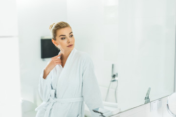 Obraz na płótnie Canvas beautiful and blonde woman in white bathrobe looking at mirror in bathroom