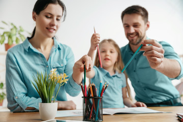 Father, mother and their daughter are smiling while spending time together. A day with family. Young happy couple with child are learning to draw. Education, studying and knowledge sharing concept.