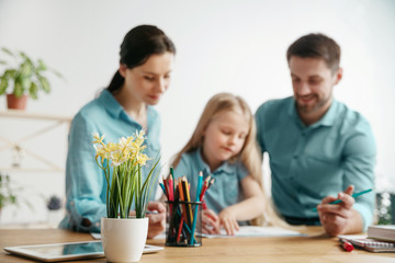 Father, mother and their daughter are smiling while spending time together. A day with family. Young happy couple with child are learning to draw. Education, studying and knowledge sharing concept.