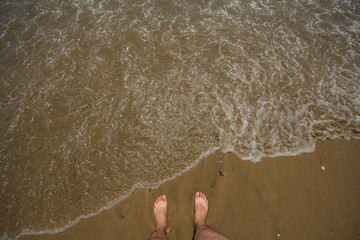 Man's legs on the sand beach and sea waves. 2 feet on the bleach sunset top view