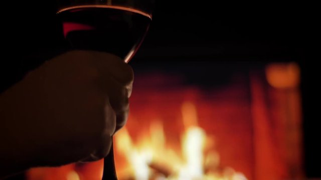 Closeup back view shoot of hands holding a glass with red wine and making a snifter with cozy warm fireplace on the background chilling and resting indoors