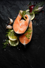 Fresh, raw salmon steaks with the addition of slices of lemon, garlic, onion, herbs and spices on a black stone background, top view. Seafood, fresh fish and ingredients for cooking