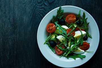 Greek salad with fresh tomatoes, arugula, eggs, olives with olive oil on a dark wood background. Healthy food. Veggie dish.