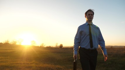 Business man with briefcase in his hand and in blue shirt with tie walks at sunset into glare of sun.