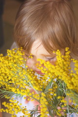 A cute little girl with light brown hair sniffs a bouquet of fragrant yellow mimosa, donated on March 8 on International Women's Day.