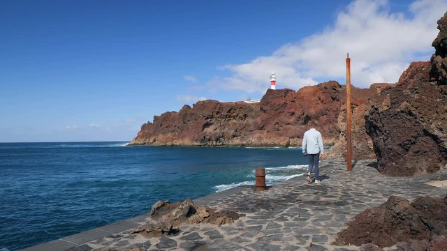 View of a tourist walking at the dock, then stops and takes a photo of the volcanic cliffs and the famous lighthouse of Teno in the Isle of Tenerife. HD cropped and Slow motion edit
