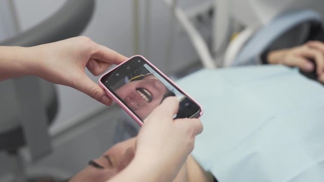 Dentist in Modern Dental Clinic Uses Her Mobile Phone Camera to Take Pictures of Teeth of Her Male Patient After Successful Treatment