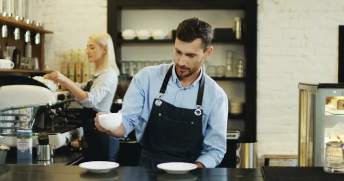 Handsome man waiter making coffee while standing at the bar and then dancing. Young blond beautiful waitress on the background.