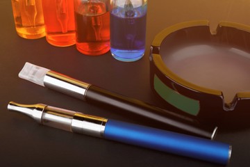 Naklejka premium Vaping devices lying near ashtray and bottles with vape liquid on black matte surface