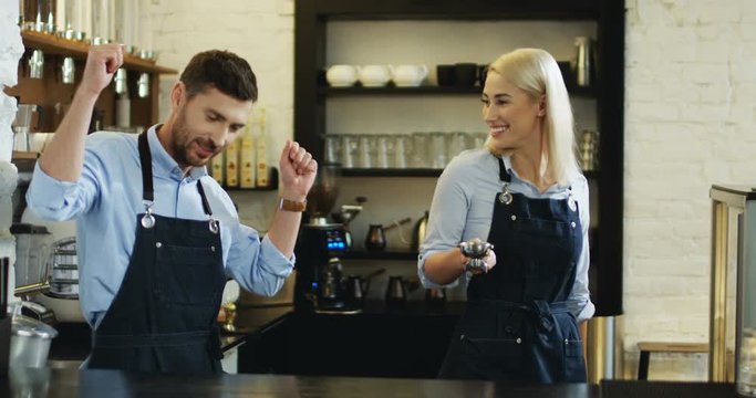 Handsome Caucasian young waiter in the apron and pretty blond waitress having fun and dancing at the bar while there are no clients.