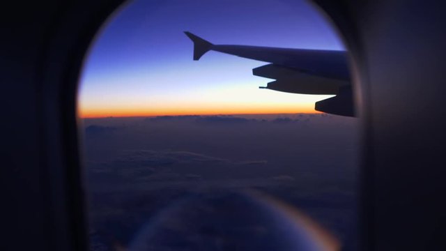 The view of the sunset blue / violet / yellow / oranges sky and the wing of the plane from the window of the porthole. The camera moves away, showing the porthole from inside the aircraft.