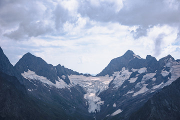 Fototapeta premium The peaks of the Caucasus Mountains in cloudy clouds and the rays of the sun through them in the outskirts of Dombai. And on the tops of snow lies.