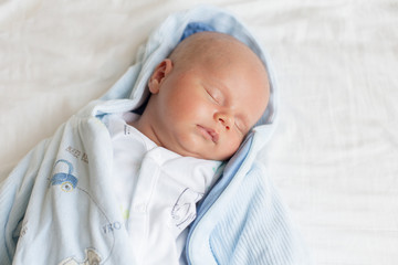 Portrait of a cute adorable baby boy of one two months old  sleeping on a bed on white shee, dreaming, wearing a light blue unfastened sleepsuit with hood.Tired after walk.