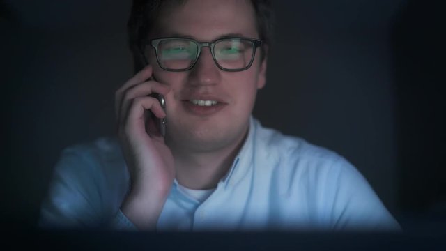 Pan shot close up of smiling man in black eyeglasses wearing blue shirt in front of laptop. Pan shot of young businessman freelancer having a call at night time working in a dark room.