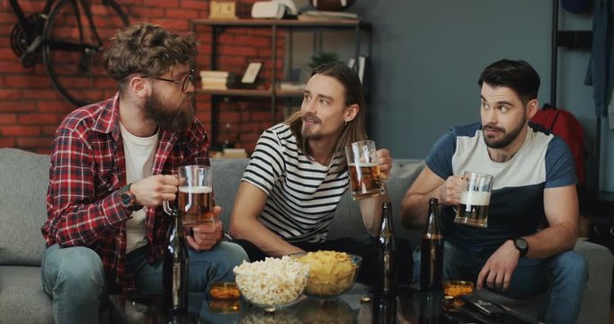 Caucasian guys drinking beer and talking on the sofa in the cozy living room during sport game on TV.