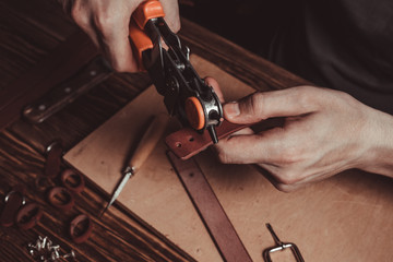 Master holding a hole punch and a piece of leather. On brown wooden table scattered with tools and accessories.