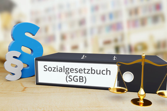 Sozialrecht. Akten-Ordner beschriftet mit dem Begriff Sozialgesetzbuch (SGB) liegt neben einer Waage und zwei Paragrafen-Zeichen auf einem Schreibtisch. B&uuml;cherregal im Hintergrund