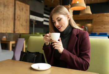 Beautiful smiling woman drinking coffee at cafe. Portrait of blonde woman in a cafeteria drinking hot cappuccino and looking at camera. Pretty woman with cup of coffee