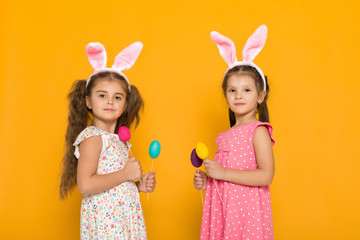 Obraz na płótnie Canvas two little girls with Easter bunny ears holding colorful eggs on yellow background. Happy easter. cute children