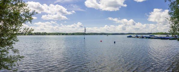 Panoramic view on the beautiful Lake Wörthsee taken from seaside. Green-blue Landscape with flag, boats, pier and plants on a clear, cloudy day. In Woerthsee, Bavaria, Starnberg, Germany.