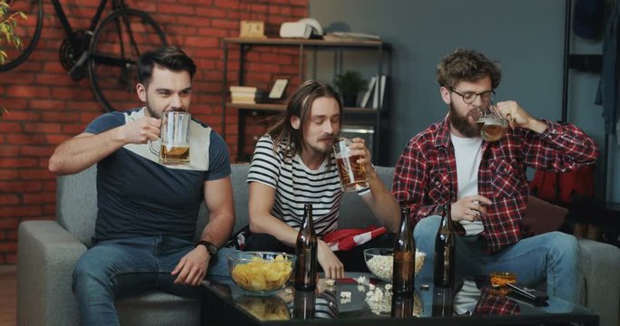 Young Caucasian Americans, male friends, sitting on the sofa in the loft styled living room and doing cheers with beers while watching sport channel on TV together.