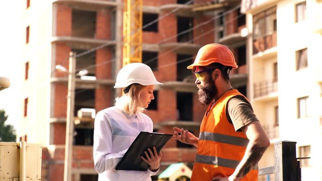 Builders communicate on the construction site. A woman in a white helmet communicates with a bearded man in an orange helmet and a protective vest on a construction site. Concept of construction.