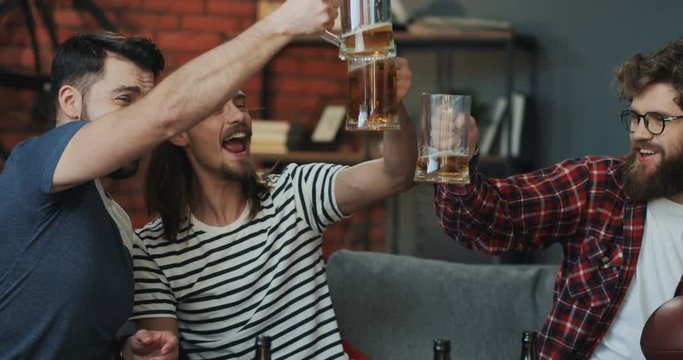 Close up of the three happy and joyful Caucasian young men, sport fans, sitting in the living room in front of TV and doing cheers with beer cheerfully.