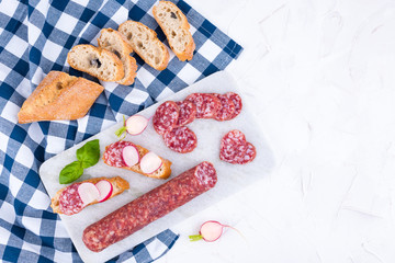 Italian salami on white board and stone background. Long sausage sliced for snacks. Free space for the text. Sandwiches with ciabatta and fresh radish. Top view