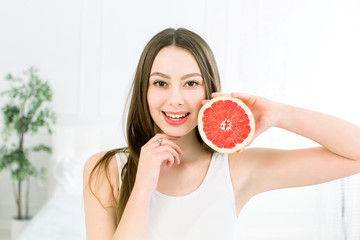 Beauty portrait of an attractive healthy woman standing isolated over light background, posing with half of juicy grapefruit near her face, and looking at camera